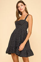 Woman wearing a black mini dress with white polka dots, a sweetheart neckline, and ruffled hem. The dress features wide elastic shoulder straps, a smocked back, and a tiered silhouette for a flattering fit.