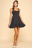 Woman wearing a black mini dress with white polka dots, a sweetheart neckline, and ruffled hem. The dress features wide elastic shoulder straps, a smocked back, and a tiered silhouette for a flattering fit.