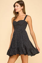 Woman wearing a black mini dress with white polka dots, a sweetheart neckline, and ruffled hem. The dress features wide elastic shoulder straps, a smocked back, and a tiered silhouette for a flattering fit.