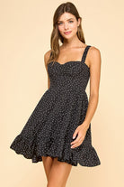 Woman wearing a black mini dress with white polka dots, a sweetheart neckline, and ruffled hem. The dress features wide elastic shoulder straps, a smocked back, and a tiered silhouette for a flattering fit.