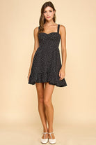Woman wearing a black mini dress with white polka dots, a sweetheart neckline, and ruffled hem. The dress features wide elastic shoulder straps, a smocked back, and a tiered silhouette for a flattering fit.