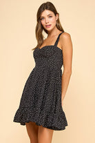 Woman wearing a black mini dress with white polka dots, a sweetheart neckline, and ruffled hem. The dress features wide elastic shoulder straps, a smocked back, and a tiered silhouette for a flattering fit.
