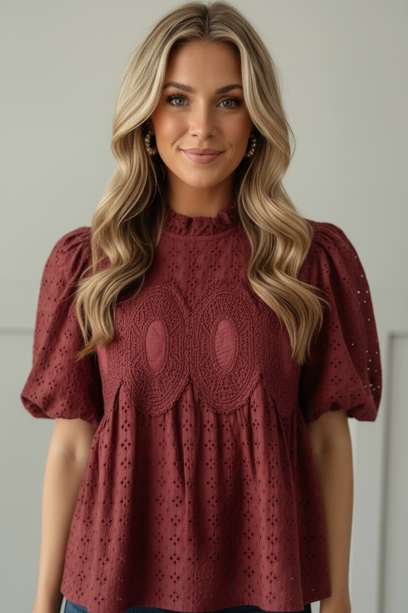 Half-body front view of cranberry burgundy eyelet puff sleeve blouse highlighting ruffled mock neckline, ornate tone-on-tone crochet medallion on bodice, and allover eyelet cotton fabric texture