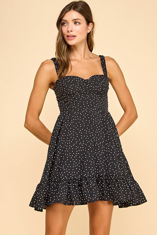 Woman wearing a black mini dress with white polka dots, a sweetheart neckline, and ruffled hem. The dress features wide elastic shoulder straps, a smocked back, and a tiered silhouette for a flattering fit.