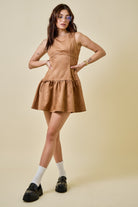 Woman wearing a camel-colored suede mini dress featuring a drop waist with bow ties, sleeveless design, and a back zipper closure. The dress has a soft, structured texture and a flattering A-line silhouette, styled for a chic fall look.