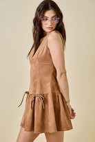 Woman wearing a camel-colored suede mini dress featuring a drop waist with bow ties, sleeveless design, and a back zipper closure. The dress has a soft, structured texture and a flattering A-line silhouette, styled for a chic fall look.
