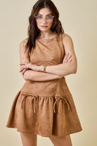 Woman wearing a camel-colored suede mini dress featuring a drop waist with bow ties, sleeveless design, and a back zipper closure. The dress has a soft, structured texture and a flattering A-line silhouette, styled for a chic fall look.