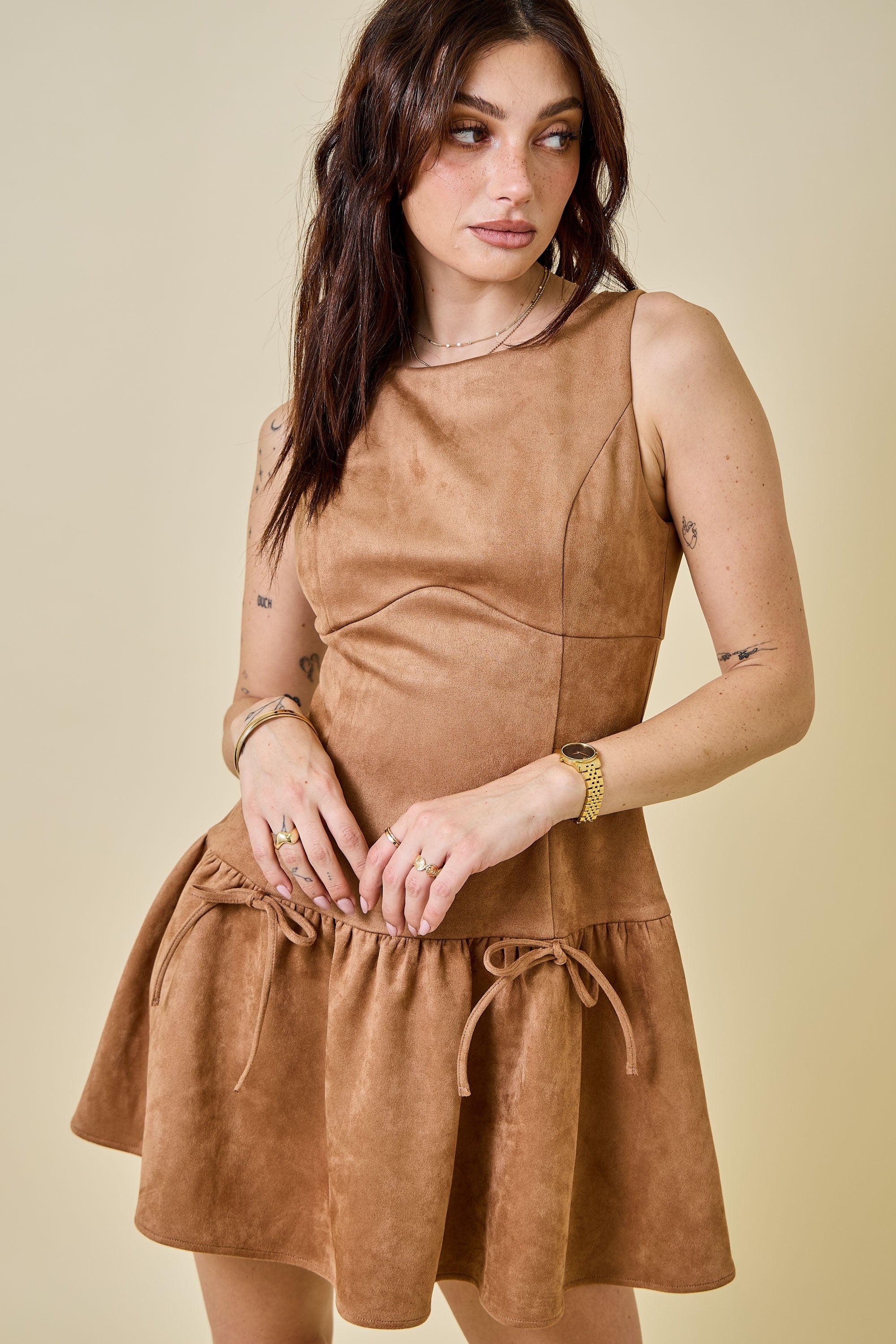 Woman wearing a camel-colored suede mini dress featuring a drop waist with bow ties, sleeveless design, and a back zipper closure. The dress has a soft, structured texture and a flattering A-line silhouette, styled for a chic fall look.