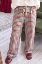 A person wearing plaid lounge pants with a white drawstring, paired with a white top and red-and-green plaid slippers, standing indoors on a light wood floor.