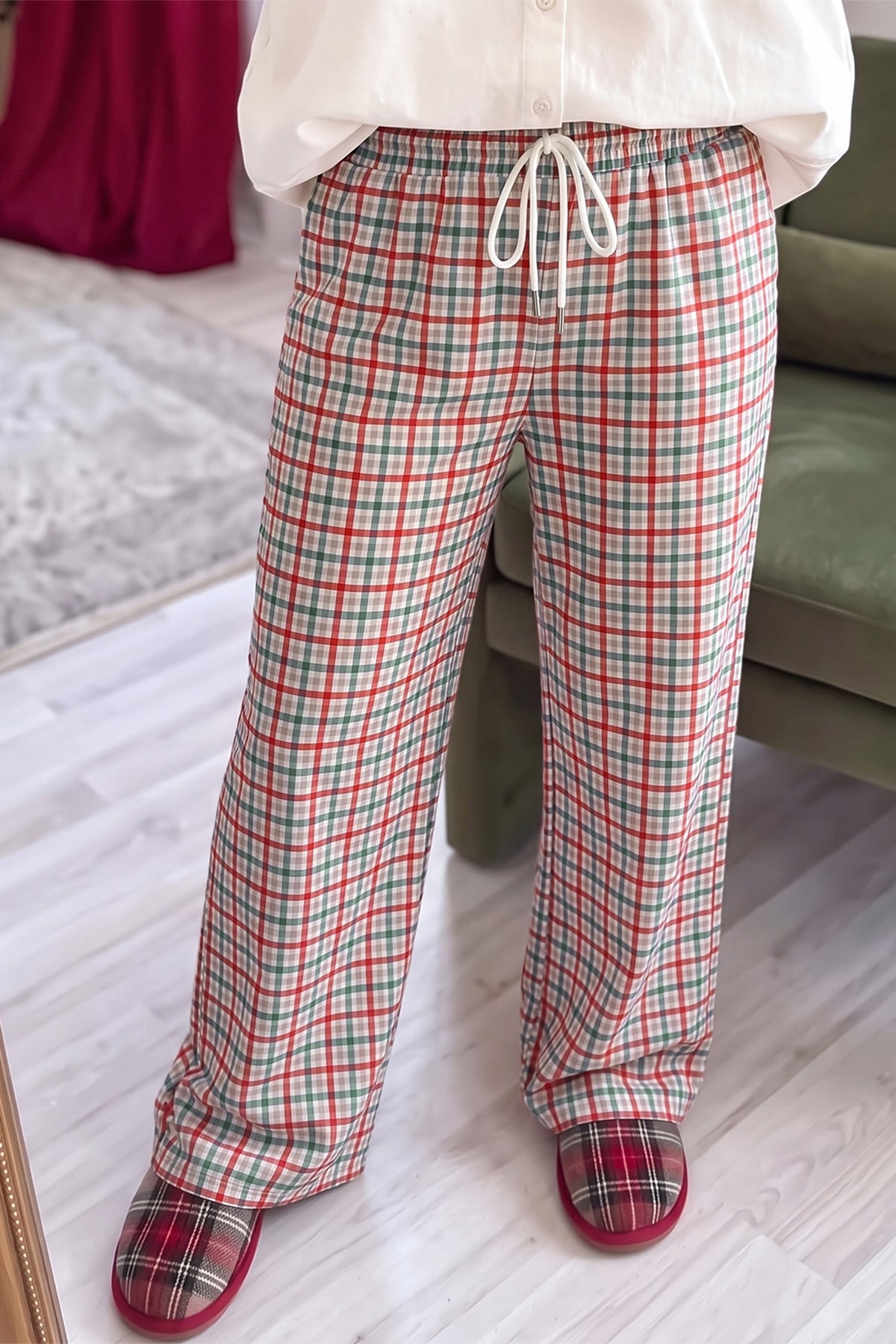 A person wearing plaid lounge pants with a white drawstring, paired with a white top and red-and-green plaid slippers, standing indoors on a light wood floor.