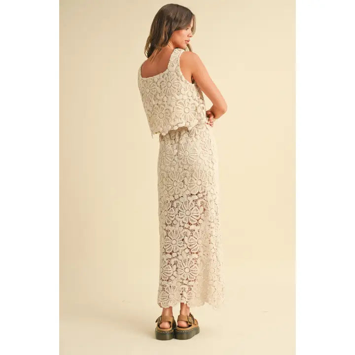 A model wearing a cream-colored crochet crop top and matching maxi skirt with a tie waist and built-in slip, styled with platform sandals against a neutral backdrop.