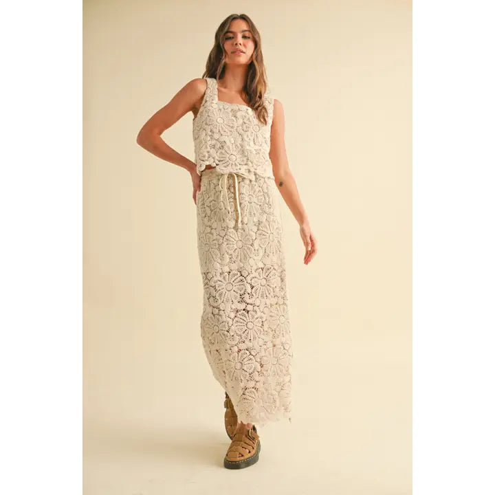 A model wearing a cream-colored crochet crop top and matching maxi skirt with a tie waist and built-in slip, styled with platform sandals against a neutral backdrop.