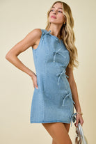 Woman wearing light blue denim sleeveless mini dress with diagonal bow tie details and exposed seam accents great for game day