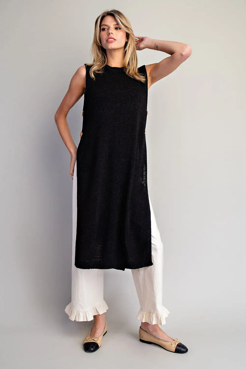 Woman wearing a black sleeveless top and white pants against a gray background