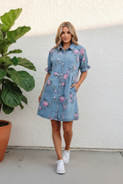 Sky blue floral denim mini shirt dress angled front view showing button-down snap closure, pointed collar, ruffle cuff sleeves, and pink rose print on light wash denim