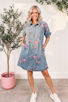 Sky blue floral print denim shirt dress front view showing snap button-down front, pointed collar, front pockets, and pink floral print, outdoor lifestyle