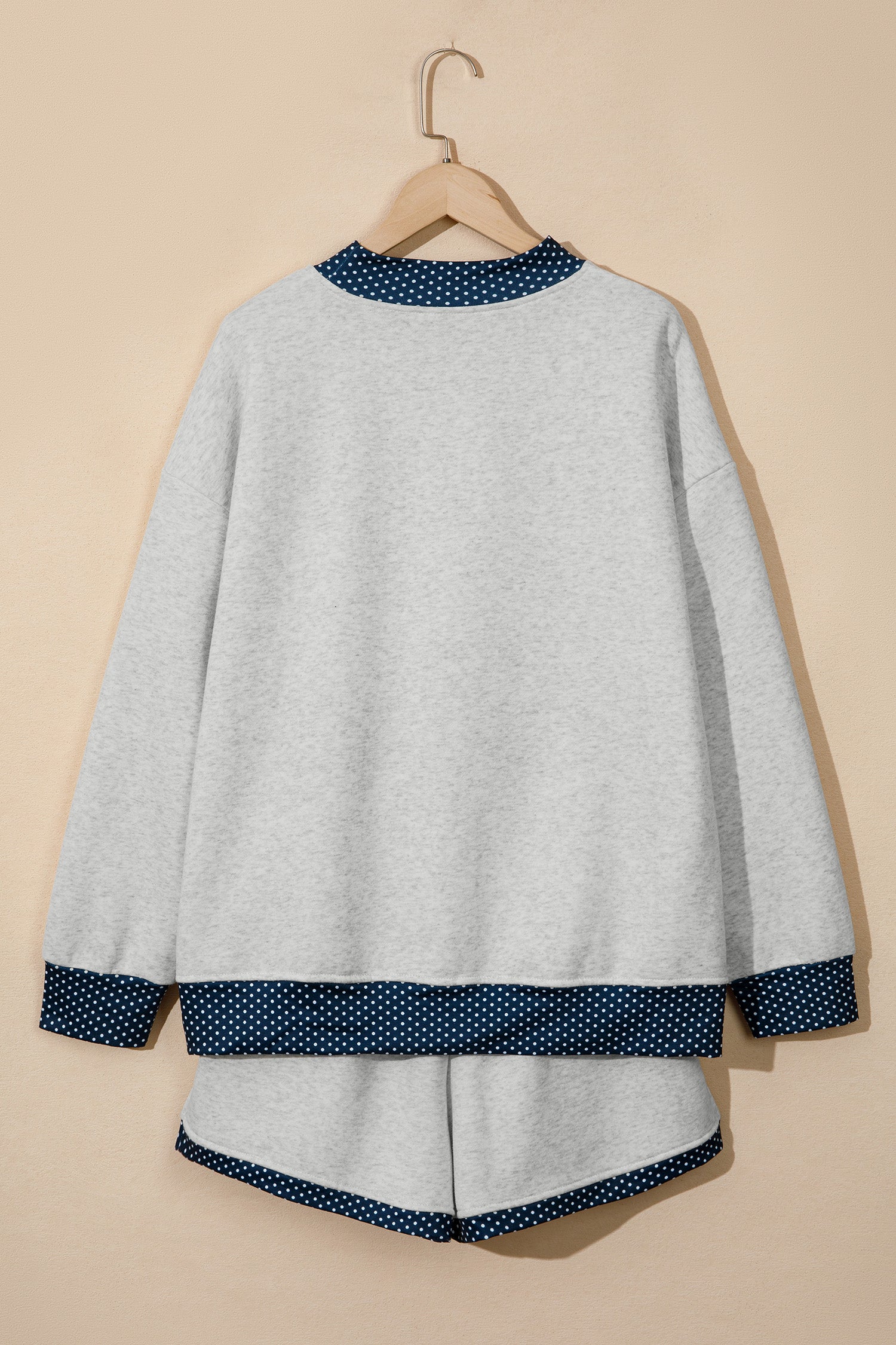 A light gray knit lounge set consisting of a long-sleeve top and matching shorts, hanging on a wooden hanger against a solid beige background. The top and shorts feature contrasting dark blue and white polka dot trim at the collar, cuffs, hem, and bottom of the shorts.