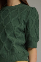 A close-up of a person wearing a dark green short-sleeve cable knit sweater with a textured diamond cable pattern and ribbed collar, cuffs, and hem.