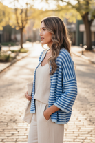 Side profile of brunette woman in blue stripe boxy knit cardigan worn open over cream linen tank top and wide-leg linen pants outdoors in natural light