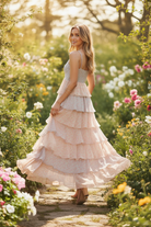 Blonde model walking in blush pink tiered ruffle maxi dress with spaghetti straps and cascading ruffle tiers in motion, sun-drenched garden path, elevated boutique lifestyle photo