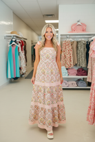 Women's pink geometric floral print tiered midi dress with pink striped patchwork bands and tie bow shoulder straps, full body front view inside boutique