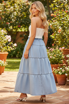Back view of Risen light wash denim high rise tiered maxi skirt showing elastic waistband panel, center back seam, and four progressively wider gathering tiers sweeping dramatically to the ankle