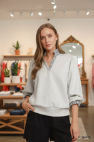 Waist-up front shot of a blonde woman wearing the Umgee heather white sweatshirt top with navy stripe ruffle collar, balloon puff sleeves, and smocked ruffle cuffs, gold hoop earrings, boutique interior