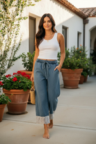 Brunette woman walking through a sun-drenched courtyard in wide-leg washed denim pants with wide floral lace ankle trim, white ribbed tank and gold jewelry — BiBi denim lace trim pants