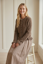 Seated smiling model in the brown Ruffle Edit cardigan, showing textured knit, a ribbed crew neckline, and wavy ruffle button placket.