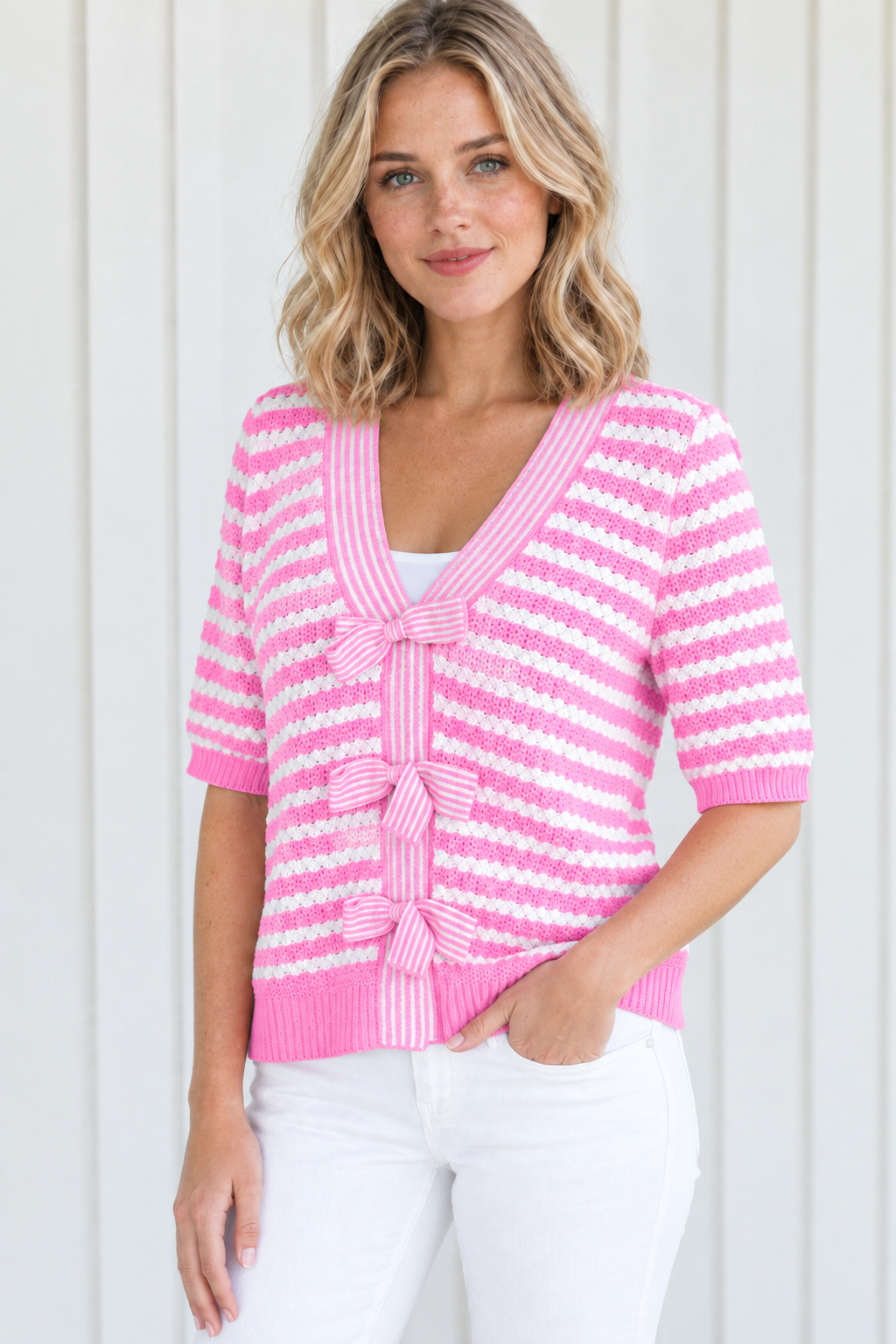 Angled front view of pink striped V-neck knit sweater with three bow ties, styled with white jeans
