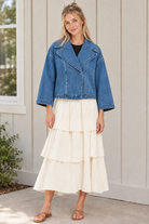 Front view of a buttoned blue cropped denim jacket with wide lapels and relaxed sleeves, worn with an ivory tiered skirt and tan sandals.
