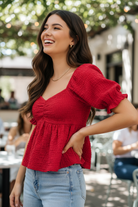 Red smocked peplum top three quarter side view — brunette model styled with wide-leg jeans outdoor cafe setting