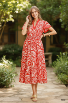 Red paisley floral print flutter sleeve midi dress full body front view, V-neckline trim, smocked waist, tiered ruffle hem, outdoor garden lifestyle photo