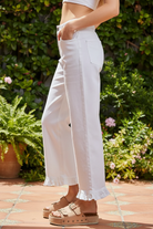 Side profile of white wide-leg mid-rise jeans with ruffled hem on blonde model, hand in pocket, outdoor patio with greenery, showing ruffle hem detail