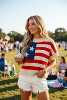 Blonde woman in a cream knit American flag sweater top with blue star canton and red cream horizontal stripes, short dolman sleeves and ribbed hem, paired with distressed white denim cutoff shorts — front full-length