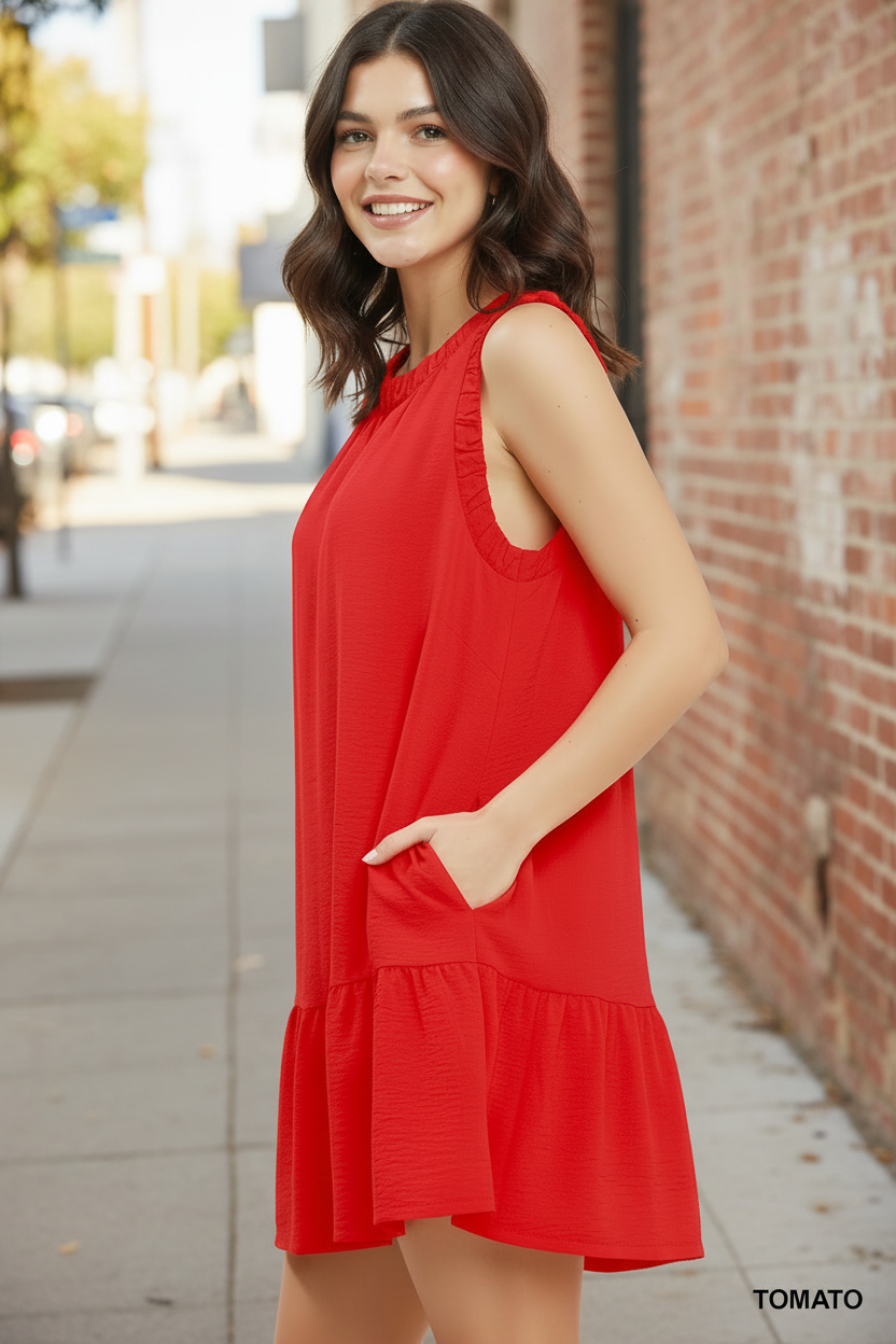 Side angle view of brunette model in Umgee tomato red sleeveless ruffle trim mini dress, hand in pocket, brick wall background
