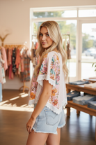 Three-quarter angle shot of a blonde woman wearing a pale pink ribbed cropped top with floral contrast flutter sleeves, eyelet lace trim strips, and scalloped lace hem, styled with light wash denim cutoff shorts in a boutique interior