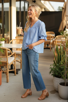 Blue gingham ruffle blouse three-quarter side view — blonde model laughing with iced coffee trendy cafe terrace barrel jeans mules