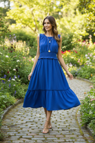 Royal blue smocked waist tiered midi dress candid walking — tiers flowing in motion brunette model garden path strappy gold sandals lifestyle