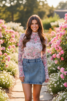 Brunette woman in a pink sheer mesh mock-neck long sleeve top with allover romantic rose floral print in pink and sage green, fitted seamless silhouette and ruched lettuce-edge cuffs, paired with denim mini shorts on a sun-drenched garden path