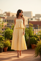 Brunette model in yellow eyelet peplum cami and midi skirt set standing on sun-drenched rooftop terrace with greenery, full body golden hour lifestyle shot