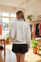 Back view of a blonde woman in the Umgee heather white french terry top with gathered back yoke, navy stripe ruffle collar, balloon puff sleeves with smocked ruffle cuffs, and ribbed hem, paired with black shorts