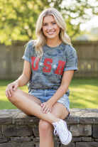 USA camo graphic tee styled with denim cutoffs — blonde model seated on steps casual lifestyle look