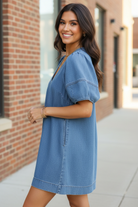 Side angle view of brunette model in Umgee light denim puff sleeve mini dress with contrast stitch V-neckline, smiling over shoulder, brick wall sidewalk