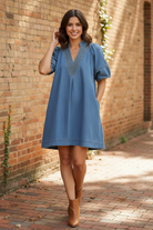 Full body front view of brunette model in Umgee light denim A-line mini dress with deep V-neckline, contrast thread stitching, and side pockets, relaxed pose on brick wall sidewalk