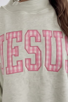 Close-up of bold JESUS pink gingham embroidered patchwork lettering on sage green oversized mock neck sweatshirt — collegiate style letters with detailed embroidery stitching and pink gingham fabric appliqué