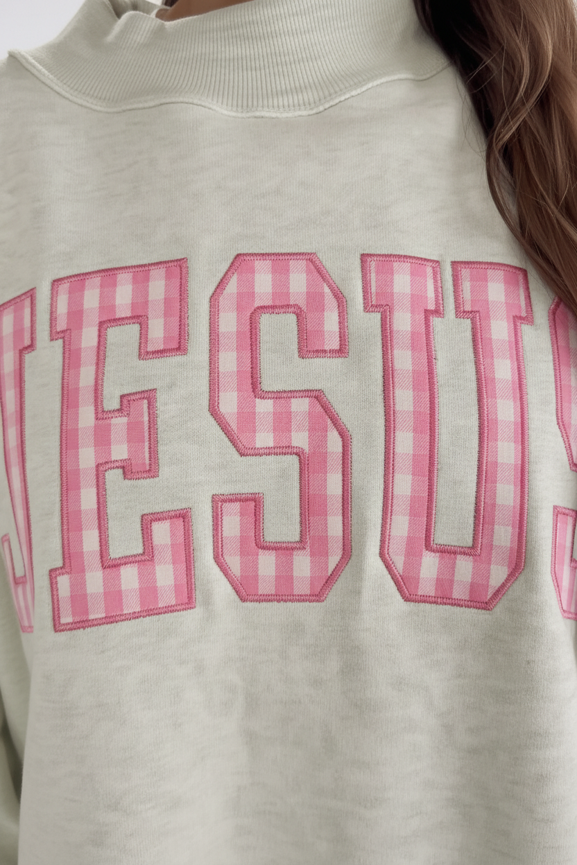 Close-up of bold JESUS pink gingham embroidered patchwork lettering on sage green oversized mock neck sweatshirt — collegiate style letters with detailed embroidery stitching and pink gingham fabric appliqué