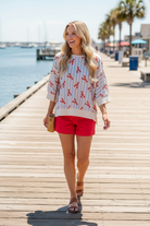 Blonde woman wearing a relaxed lobster and stripe print top with waffle knit short sleeves and red shorts, walking along a sun-drenched waterfront boardwalk — patriotic summer top