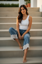 Brunette woman sitting on sunlit stairs showing full lace hem detail on wide-leg washed denim pants, white ribbed tank tucked in, gold jewelry — BiBi floral lace trim denim pants