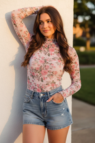 Brunette woman in a pink floral rose print sheer mesh mock-neck top with ruched lettuce-edge cuffs, arm raised candid pose against a white wall in golden afternoon light, paired with light wash denim mini shorts
